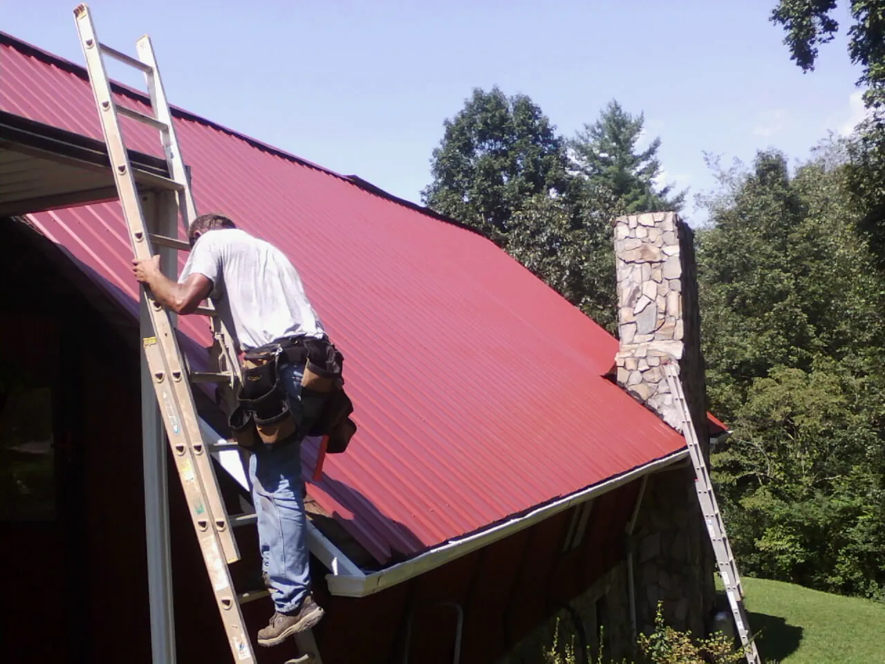 Roofer on a ladder beside a red metal roof and stone chimney — Soffit & Fascia Repair by Roofing Brothers in Brinklow